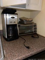 Photo of a stainless steel Cuisinart coffee maker and a Black+Decker toaster oven on a countertop with trays stacked on the oven, power cords visible.