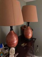 Photo of two matching ceramic base table lamps with beige shades on a dresser with surrounding items.