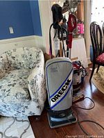 Full view of the Oreck upright vacuum cleaner showing the fabric dust bag, handle, and base on wood floor next to a chair.