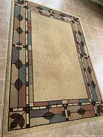 Full view of the rectangular area rug showing beige center and patterned border with leaf motifs on tiled floor.