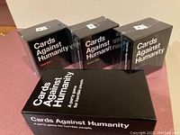 Four sealed boxed sets of Cards Against Humanity including Red Box, Green Box, Blue Box, and original black box, displayed on a table.