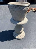Front view of the cement planter on the decorative stand showing the diamond lattice and rope-like designs on the planter and leaf carvings on the stand base and top.