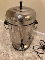 Front view of stainless steel coffee urn with black handles and lid knob, showing overall appearance.
