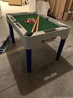 Photo of the child's 4-in-1 game table showing green felt pool playing surface, two pool cues on the table, blue legs, and pool balls in triangular rack.