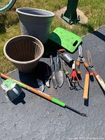 Overview of assorted gardening tools and plastic planter pots placed outdoors on paved surface and grass.