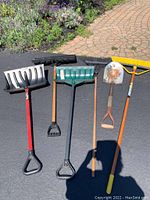 Photo showing 3 snow shovels with plastic blades and D-shaped handles, one short spade with wooden handle and rusted metal blade, and two push brooms with wooden handles and stiff bristles.