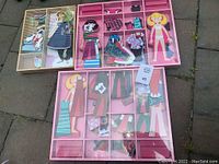 Two sets of Melissa & Doug Abby & Emma magnetic wooden dress-up dolls with various magnetic clothes and accessories arranged in segmented trays.