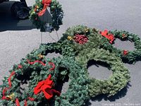 Photo showing six oversized artificial Christmas wreaths and one smaller wreath placed on pavement, decorated with red bows, ribbons, and some with berries.