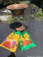 Large metal plant stand with wide bowl top, smaller metal stand with cylindrical holder, and two partially full bags of potting soil on paved surface outdoors