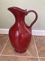 Red ceramic jug with curved handle and wide pouring spout on tiled floor, side view.