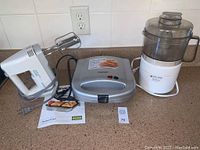 Three kitchen appliances displayed on countertop: Black and Decker hand mixer, Cuisinart sandwich grill, Black and Decker food chopper, with sandwich grill manual and lot number card
