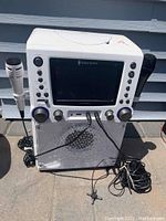 White Singing Machine karaoke player with two microphones attached, front view showing screen and controls.