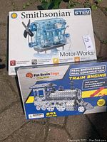 Photo showing both Smithsonian Motor-Works STEM kit and Fat Brain Toys Train Engine construction kit boxes outdoors on a stone surface.