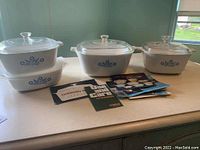 Full set of three CorningWare Cornflower Blue baking dishes stacked and displayed with lids and instruction booklets on table.