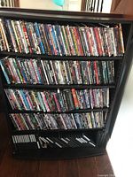 Full view of black wooden storage shelf filled with assorted DVDs vertically arranged on three shelves, some empty slots at bottom.