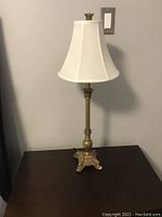 Full view of one bedside table lamp with white tapered lampshade and gold ornate base standing on a dark wooden surface against a gray wall.