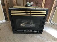 Front view of black and gold gas fireplace insert, showing window with decorative gold horizontal trim and logs inside.