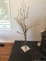 Gold-colored tree sculpture with bare branches mounted on a square mirror base, placed on wooden floor near window and wall.