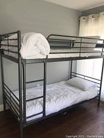 Front view of grey metal single bunk beds with mattresses and a white comforter and pillow on bottom bunk and rolled bedding on top.