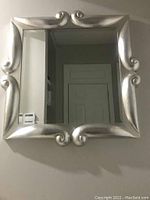 Front view of square mirror with silver wood frame showing decorative scroll details and clear reflective surface.