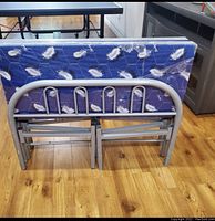Folded bed frame showing the metal tubular headboard and footboard and folded mattress with blue feather patterned fabric cover.