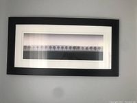 Photo showing black rectangular frame with white matting containing a panoramic grayscale image of a row of trees.