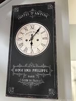 Front view of the black rectangular wall clock with round white clock face featuring Roman numerals and Parisian text design.
