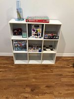 White 3x3 cubby cube open storage cabinet holding board games, against a white wall on wooden floor