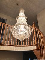 Large, multi-tier chandelier hanging from ceiling in stairwell area, showing clear imitation crystals and gold-tone metal frame with chain.