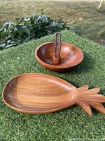 Wooden nut bowl with inset metal nutcracker and six nut picks; pineapple-shaped wooden catchall tray on grass outdoors.