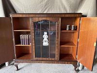 Open front view showing two wooden side doors open, middle door with glass and internal glass shelves, containing a white sculptural object, books on left side shelves, two mugs on right side shelves.