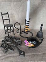 Overview of set showing all six cast iron items on beige fabric background: trivet, chair, candle holder with candle, bell, snuffer, and bird-shaped match holder with matches.