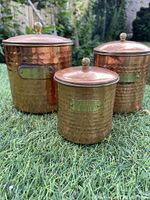 Photo of three hammered copper canisters with brass knobs on lids, placed on grass outdoors, showing varying sizes