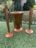 Photo showing set of three copper vases: two slender bud vases and one larger flared vase, placed outdoors on a table with grass in background.