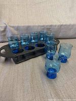 Eight blue vintage glasses placed in a black wooden holder tray with cut-out slots for each glass.