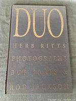 Front cover of the photobook featuring title 'DUO' and artists' names in gold embossing on dark linen fabric.