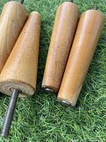 Four tapered wooden table legs with metal screws attached, shown on a grass background. Two legs are shorter (approximately 6 inches), two are longer (approximately 14 inches).
