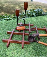 Photo of two wooden lattice trivets, a two-arm candle holder with brass collars on wooden candle cups, and a wooden handled metal nutcracker, arranged on grass.