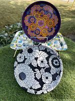 Three retro tin trays arranged outdoors. Two round trays with floral designs and one rectangular tray with colorful pea pod pattern visible.
