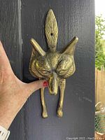 Photo showing entire fox-shaped brass door knocker mounted on dark wood door, hand nearby for scale.