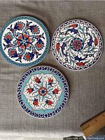 Three round Turkish ceramic tiles with colorful floral patterns laid on a gray fabric backdrop with a ruler showing 6-inch size.