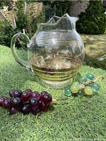Photo showing clear glass ball pitcher with gold horizontal stripes and two resin grape clusters on wire, placed on green grass outdoors.