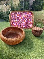 Overview of the three items on grass: the vinyl-lined wicker tray with colorful circular pattern, the round woven bamboo basket, and the smaller lidded basket with handle.