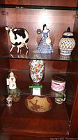 Glass cabinet shelf with various collectibles including ceramic Asian style vase, candle votive, Bavarian mug, and figurines.