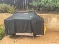 Full view of the covered BBQ grill placed outdoors on patio with surrounding plants and fencing.