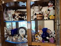 Wide view of wooden display cabinet shelves containing assorted cow collectibles including cookie containers, figurines, plates, and small vases.