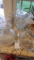 Seven various clear glass vases displayed on a kitchen counter with granite surface, positioned near a window and next to a plant.