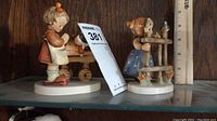 Photo showing both Hummel figurines side by side on a wooden shelf with a ruler for scale.