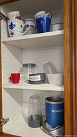 Shelves inside a white cabinet holding mugs, plastic containers, and parts of the NutriBullet Magic Bullet blender set.