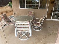 Wide view of outdoor round glass table and five chairs with floral cushions on patio.